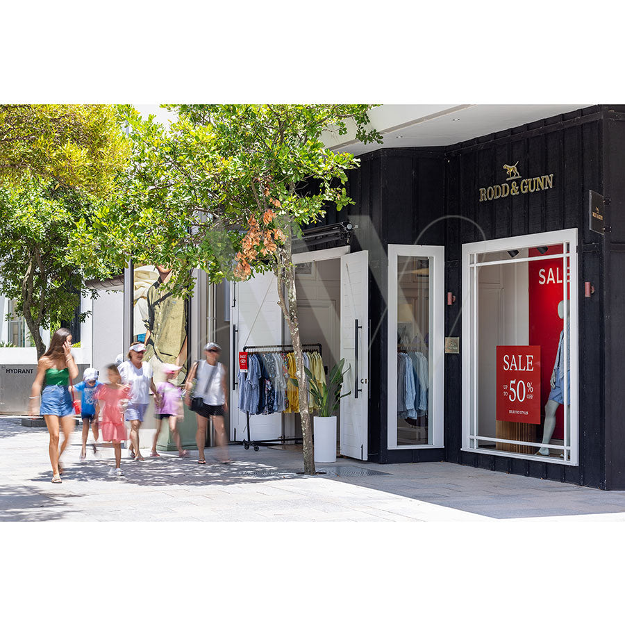 In Hastings Street Image 5413 from AWP Image Library, a lively scene shows people passing by a Rodd & Gunn store with an open entrance, clothing racks visible inside, and a large red sale sign offering up to 50% off. Lush green trees offer shade to the bustling sidewalk.