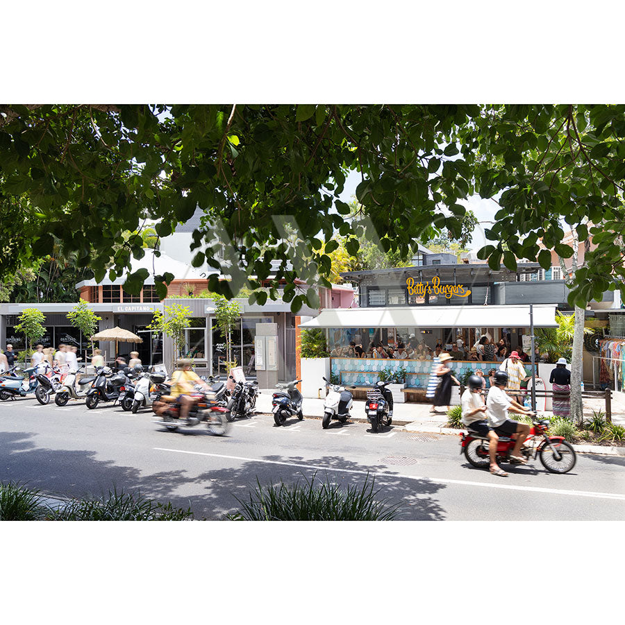The Hastings Street Image 5593 by AWP Image Library captures a vibrant scene with motorbikes cruising past a contemporary burger joint, large trees offering shade, and pedestrians strolling by, creating a bright and bustling atmosphere.