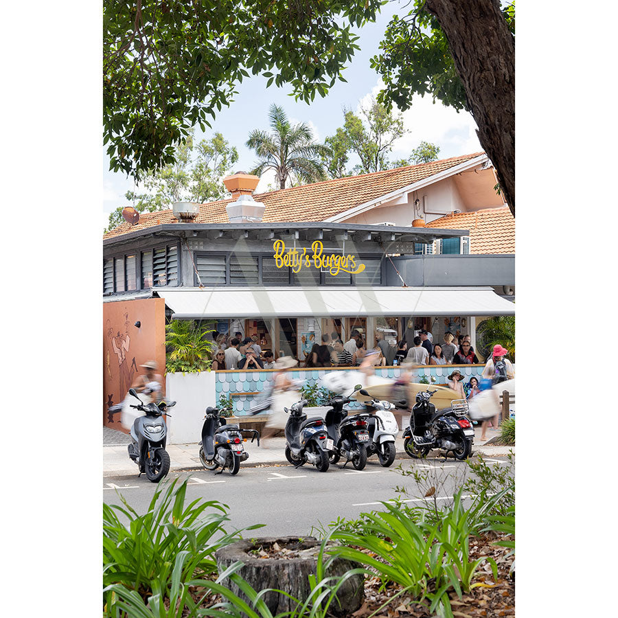 Hastings Street Image 5695 by AWP Image Library captures a lively street scene with scooters parked by Bettys Burgers, people dining on the outdoor patio beneath a white awning amid greenery near El Capitano.