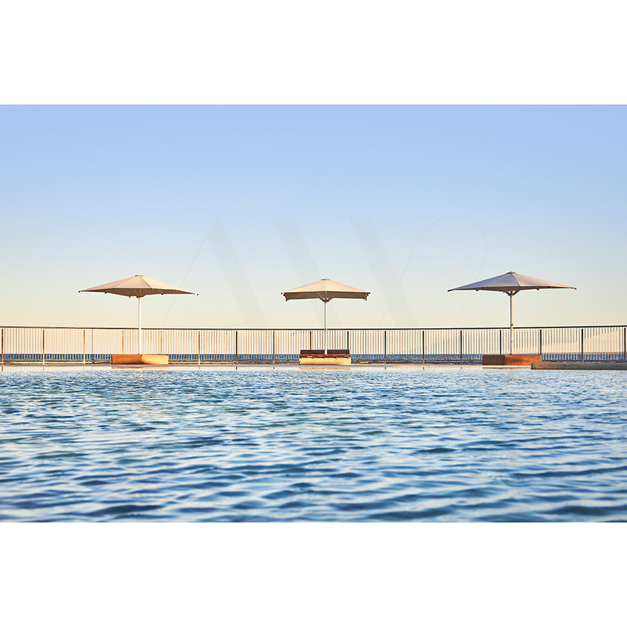 Kings Beach Pool Image 8241 by AWP Image Library captures a poolside view with two umbrellas over sun loungers by an infinity pool. The calm water mirrors the clear blue sky, while a fence divides the pool area from the horizon.