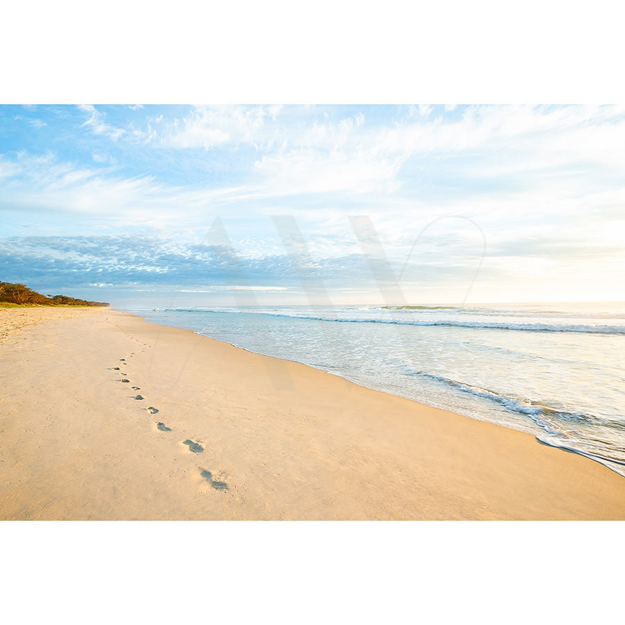 The Kingscliffe Beach Image 0606 by AWP Image Library captures footprints trailing into the distance on wet sand, gentle waves lapping ashore under a partly cloudy blue sky with sunlight casting a warm glow over the serene beach scene.