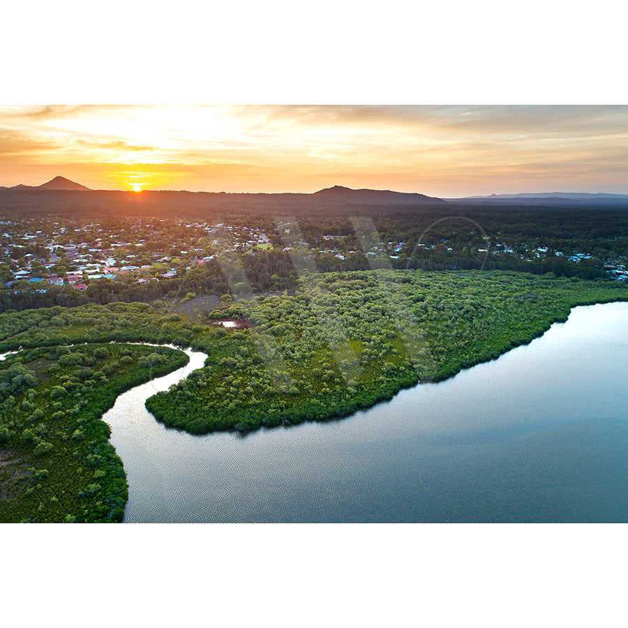 Lake Doonella Image 0072 by AWP Image Library captures a drone’s aerial view of a winding river near Lake Doonella, bordered by lush greenery, with a town and distant mountains under a vibrant sunset sky.