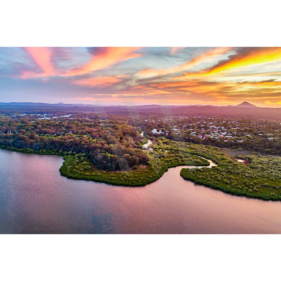 Lake Doonella Image 0100 by AWP Image Library captures an aerial view of a scenic sunset landscape with a winding river bordered by lush greenery, a small town nestled among trees, and colorful clouds reflecting orange, pink, and purple hues over the water.