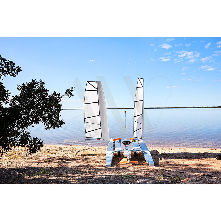 The Lake Cootharaba Image 3099 by AWP Image Library portrays a catamaran with white sails on the sandy shore, under a clear sky with few clouds and a leafy tree, capturing the natural beauty near the Noosa Everglades.