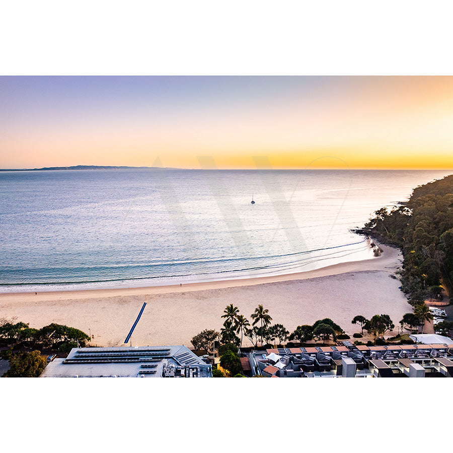 The Main Beach Drone Image 3473 by AWP Image Library features a tranquil beach at sunset with tree-lined shores, a few buildings, and a calm ocean stretching to the horizon where a boat is visible under an orange to purple sky.