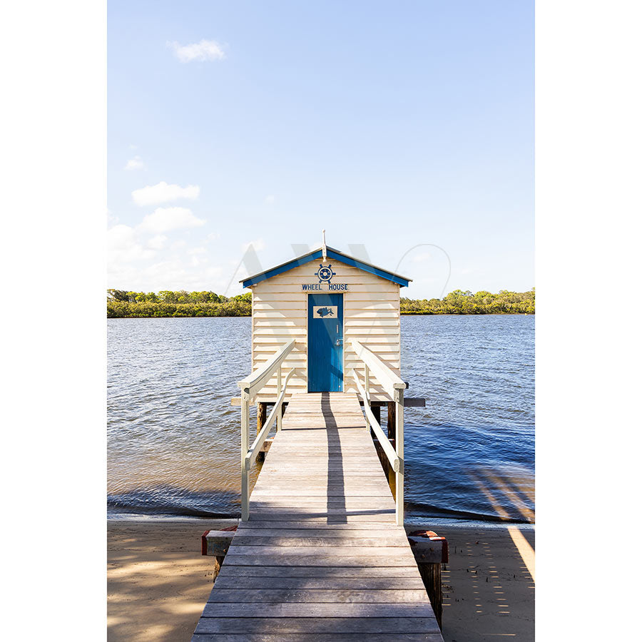 The Maroochy Boat House Image 2185 by AWP Image Library portrays a wooden boathouse labeled BOAT HOUSE above its blue door, situated at the end of a scenic boardwalk over calm waters, framed by lush greenery near Coolum Beach under a clear sky.