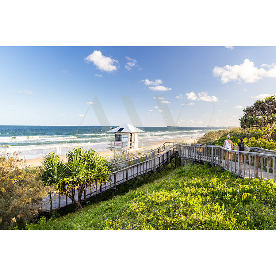The Mount Coolum Image 5776 from AWP Image Library captures a coastal scene with a wooden boardwalk leading to the beach, featuring a small lifeguard tower by the shore. Two people sit on the railing enjoying the calm ocean and partly cloudy sky, surrounded by lush greenery.