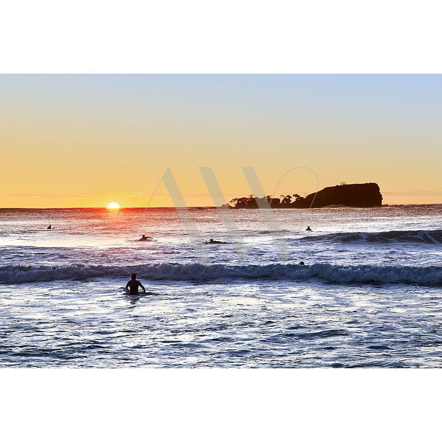 Mudjimba Surfers Image 5223 by AWP Image Library captures surfers riding waves at sunrise on Mudjimba Beach, with the sun casting a warm glow and Old Woman Islands silhouette enhancing the serene scene.