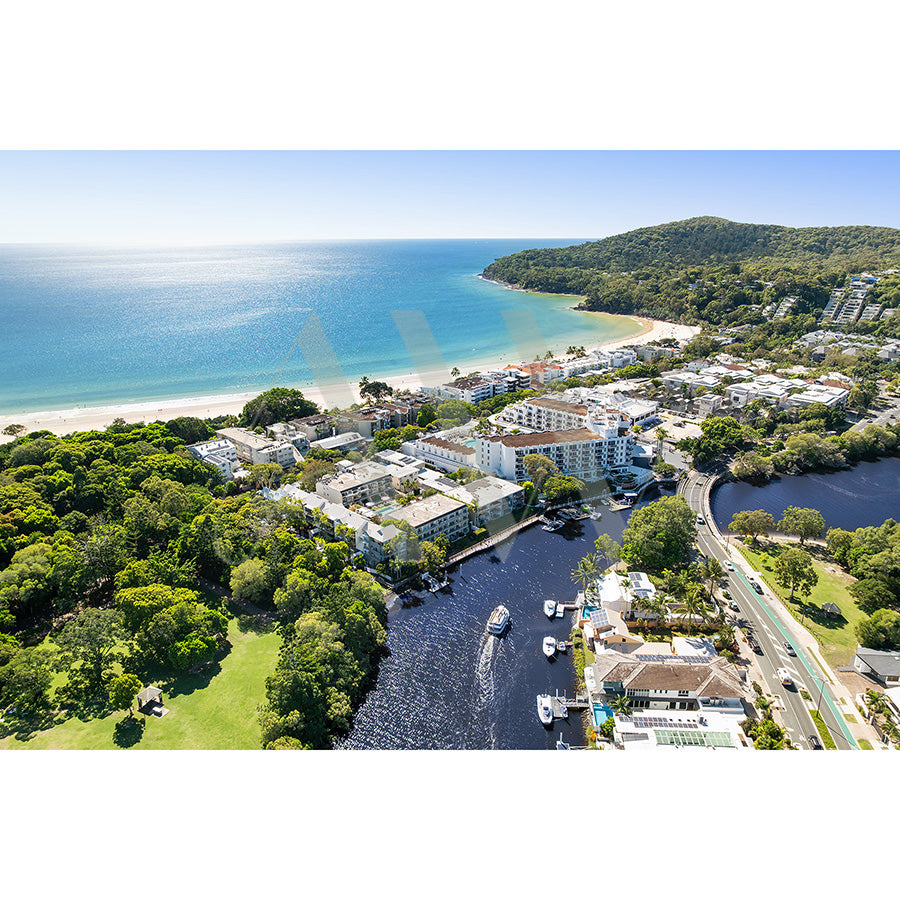 Noosa Drone Image 2359 by AWP Image Library captures a coastal town with white buildings, lush greenery, boats on the Noosa River, sandy beach meeting turquoise waters, and hills under a clear blue sky.