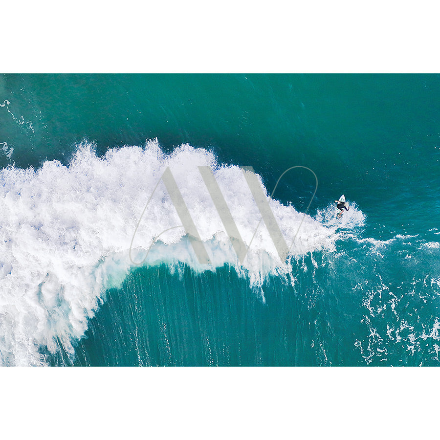 The Noosa Drone Image 0028 by AWP Image Library captures a surfer riding a turquoise wave, with white surf trailing dynamically behind. The calm, expansive ocean contrasts with the waves energy.