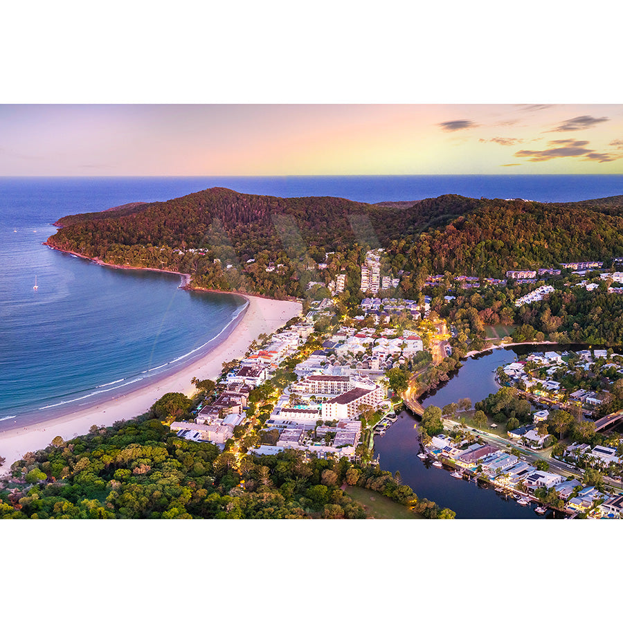 The Noosa Drone Image 0393 by AWP Image Library captures a stunning aerial view of a coastal town, featuring Noosa Main Beachs curved sandy shore and calm blue waters. Surrounded by forested hills and nestled greenery, Hastings Street lines a river under the twilight skys warm hues.