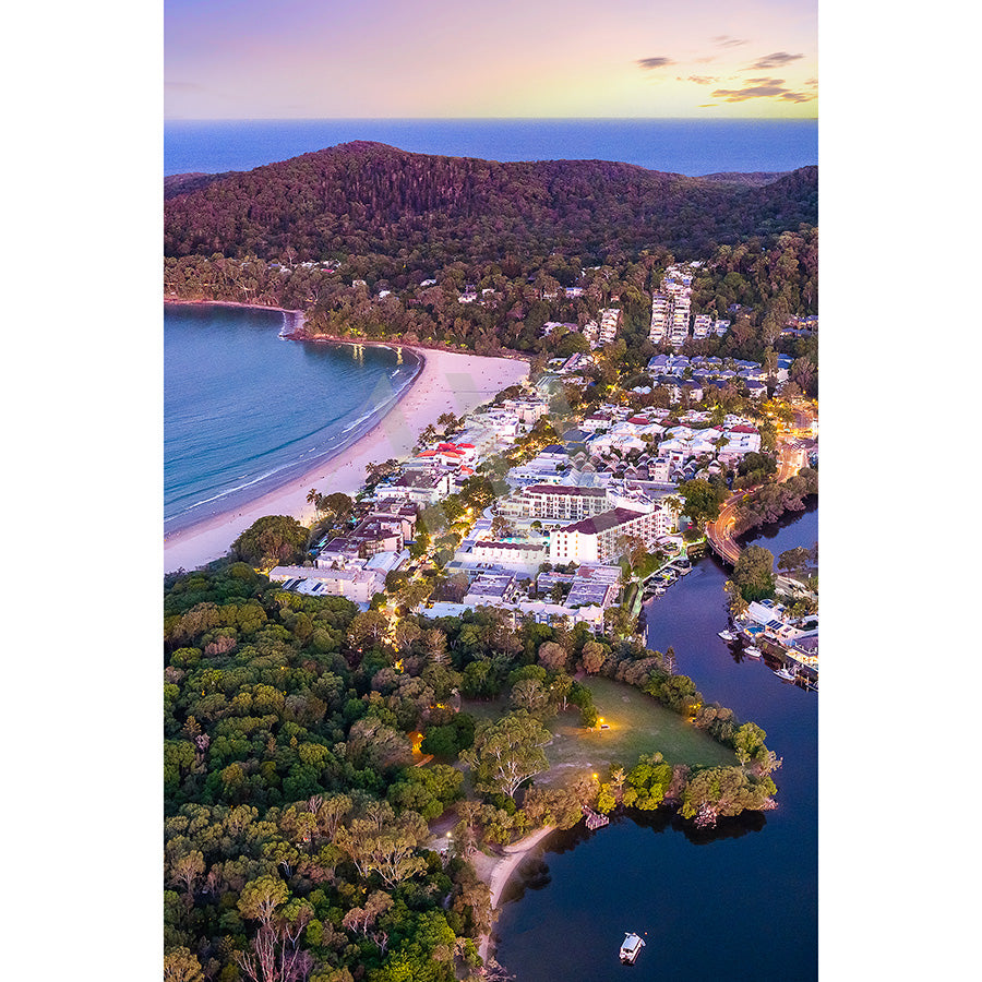 Experience Noosa Drone Image 0420 by AWP Image Library: an aerial view of a coastal town with sandy beaches, lush hills, and a serene bay at sunset. White buildings peek through trees near a calm river with boats, creating a picturesque and tranquil scene.