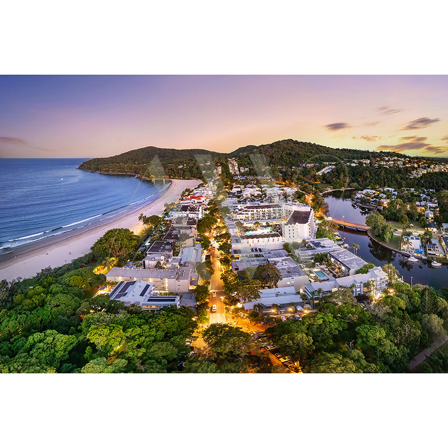 The Noosa Drone Image 0489 from AWP Image Library captures a coastal town between a lush forest and sandy beach, illuminated by lights at sunset. Hills frame the scene, and a calm body of water is visible, all under a warm glow.