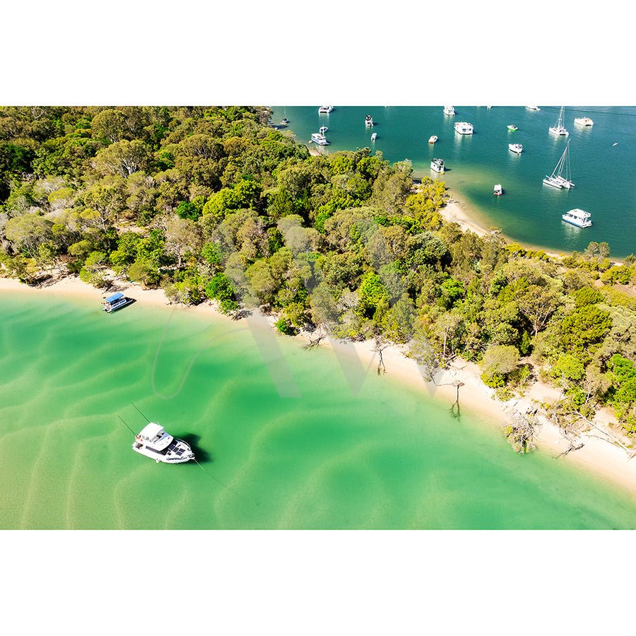 The Noosa Drone Image 0788 from AWP Image Library captures a lush, forested shoreline along the serene Noosa River, with boats moored nearby. The scene boasts vibrant greenery under clear skies and sparkling turquoise waters, epitomizing a tranquil tropical atmosphere.