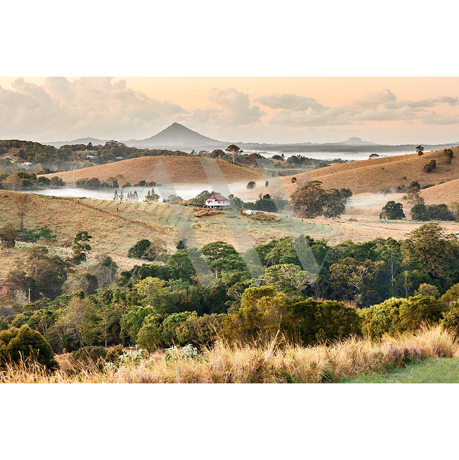 Noosa Hinterland Image 2372 from AWP Image Library portrays a peaceful rural setting with a lone house amidst green and golden fields at sunrise. Mist drapes over the hilly landscape, while distant mountains peek under a cloudy sky.