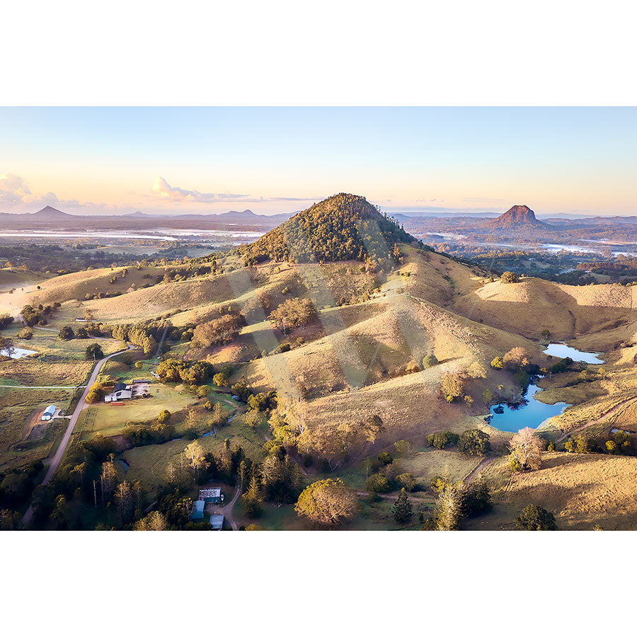 The Noosa Hinterland Drone Image 0650 by AWP Image Library captures a breathtaking aerial view of hilly landscapes at sunrise, with golden light illuminating grassy hills and trees. Small lakes and scattered houses are visible across the vast expanse.