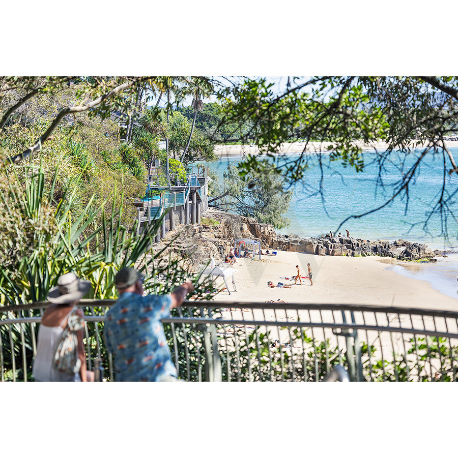Noosa Image 5083 from AWP Image Library shows two people on a railing overlooking a sandy beach at Noosa National Park, with sunbathers, swimmers, pandanus trees, rocks, lush greenery, and calm blue ocean waters in the background.
