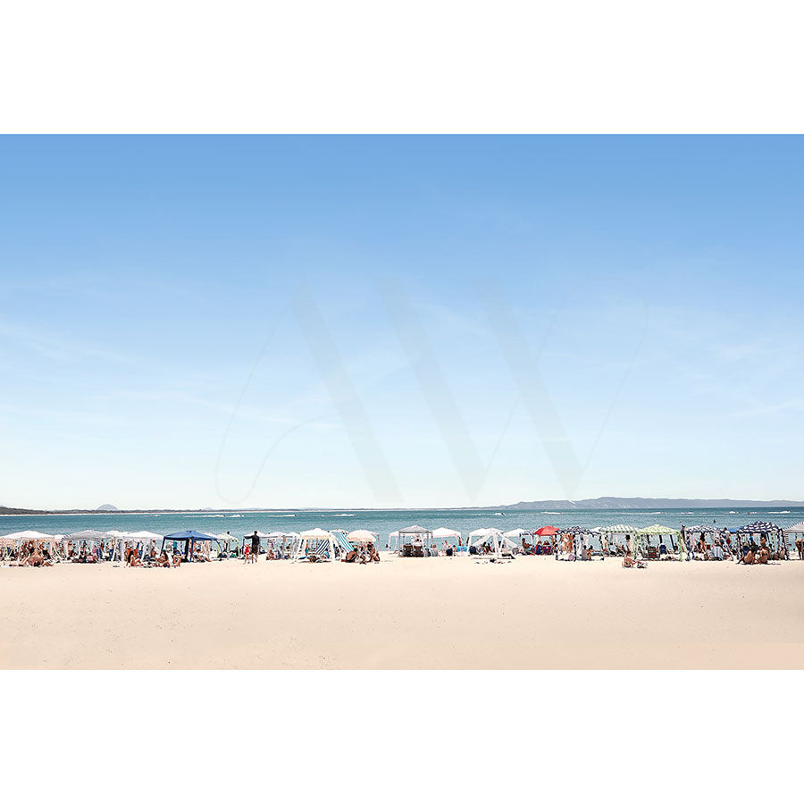 The Noosa Main Beach Image 1095 from AWP Image Library captures a lively beach scene with many sunbathers under white umbrellas. The sandy shore stretches in the foreground, while the calm blue sea extends to the horizon. A clear sky amplifies the summery vibe.