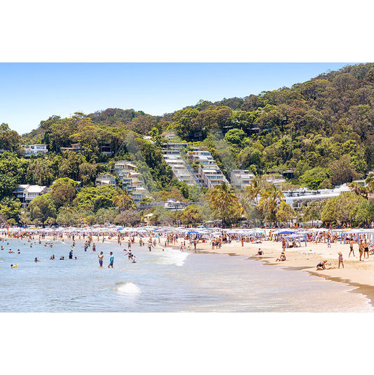 AWP Image Librarys Noosa Main Beach Image 4695 captures a sunny beach day with gentle waves, lush green hills, scattered beachgoers, umbrellas dotting the sandy shore, and hillside houses peeking through the trees.