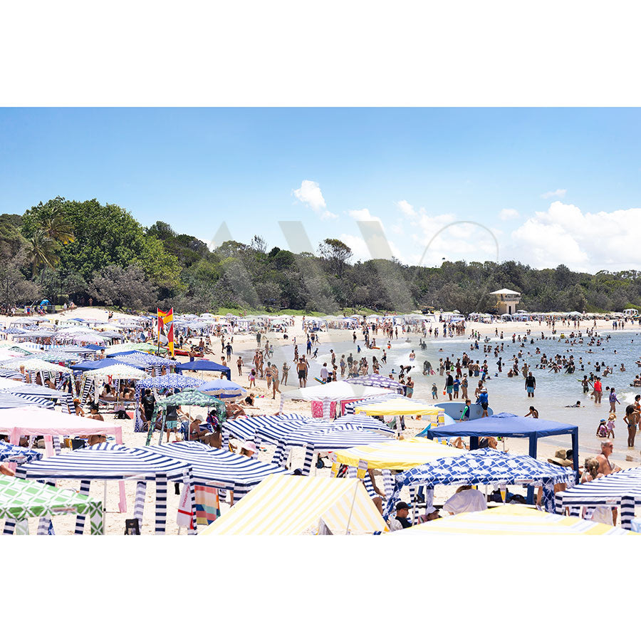 The Noosa Main Beach Image 4798 from AWP Image Library captures a vibrant beach with people swimming and relaxing under striped umbrellas, set against calm ocean waters and a tree-lined shore beneath a clear blue sky.