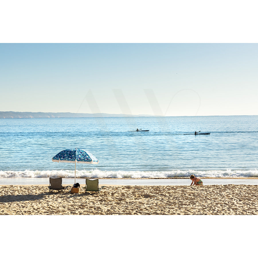 An image titled Noosa Main Beach Image 4874 from AWP Image Library depicts a beach scene with two empty chairs and a blue umbrella. Nearby, someone plays in the sand, while two kayakers paddle on the tranquil ocean under a clear blue sky.