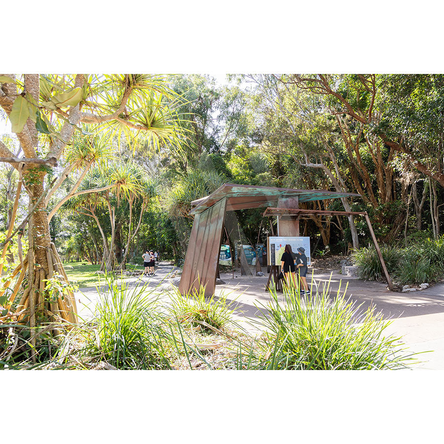 Noosa National Park Image 5848 by AWP Image Library captures a modern, angular metal structure along a sunlit path, surrounded by lush greenery and tropical plants, with people enjoying the vibrant park setting.