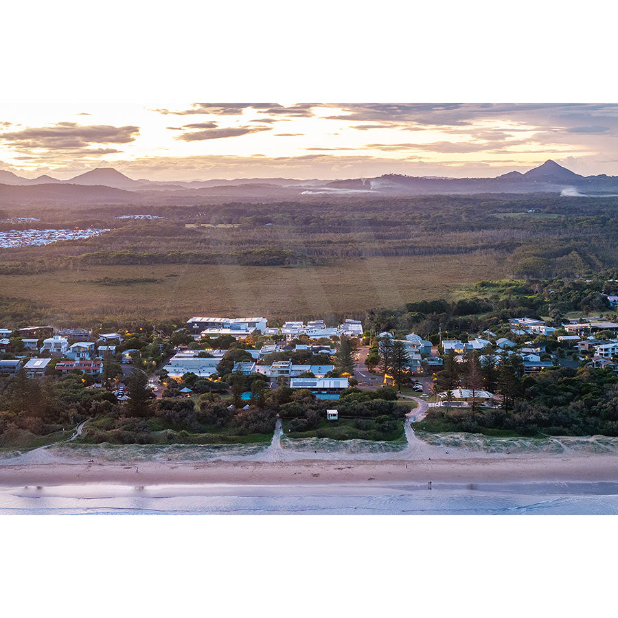 Peregian Beach Drone Image 0243 by AWP Image Library captures an aerial view of a coastal town at sunset, featuring tree-surrounded houses, a beach in the foreground, and fields with distant mountains beneath a cloudy sky on the horizon.