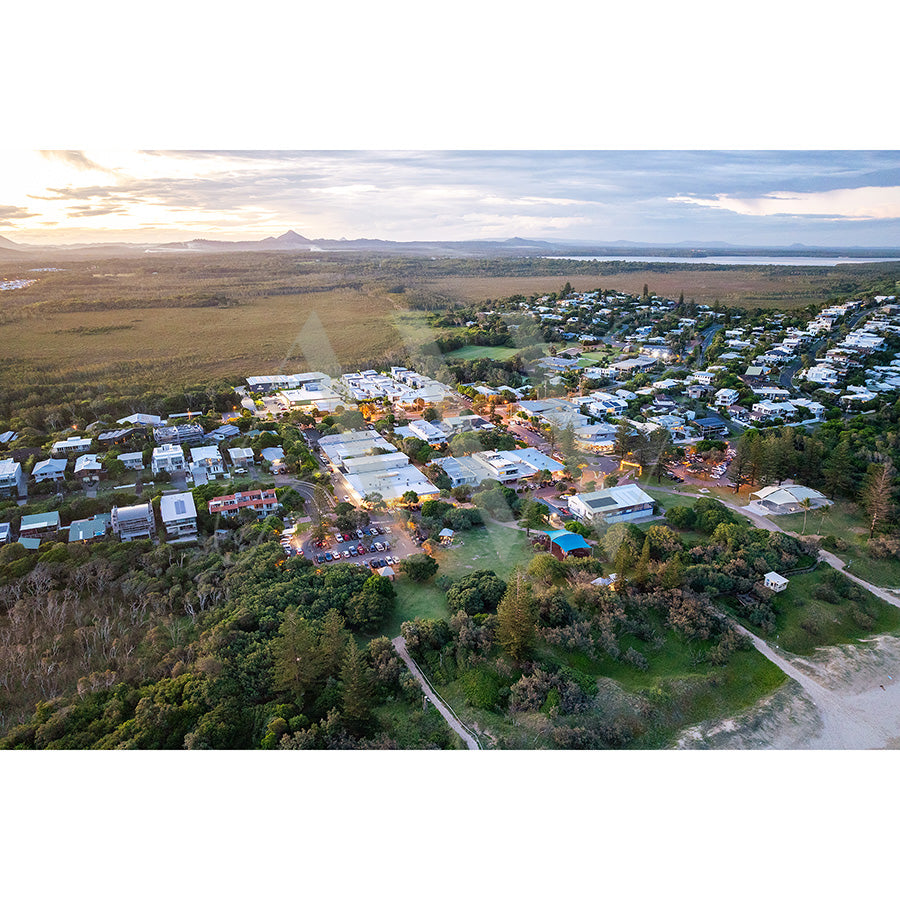 The Peregian Beach Drone Image 0275 by AWP Image Library captures an aerial view of a coastal town, houses clustered among greenery and bordered by beaches and forest. The horizon reveals a sunlit sky, clouds, and distant mountains.