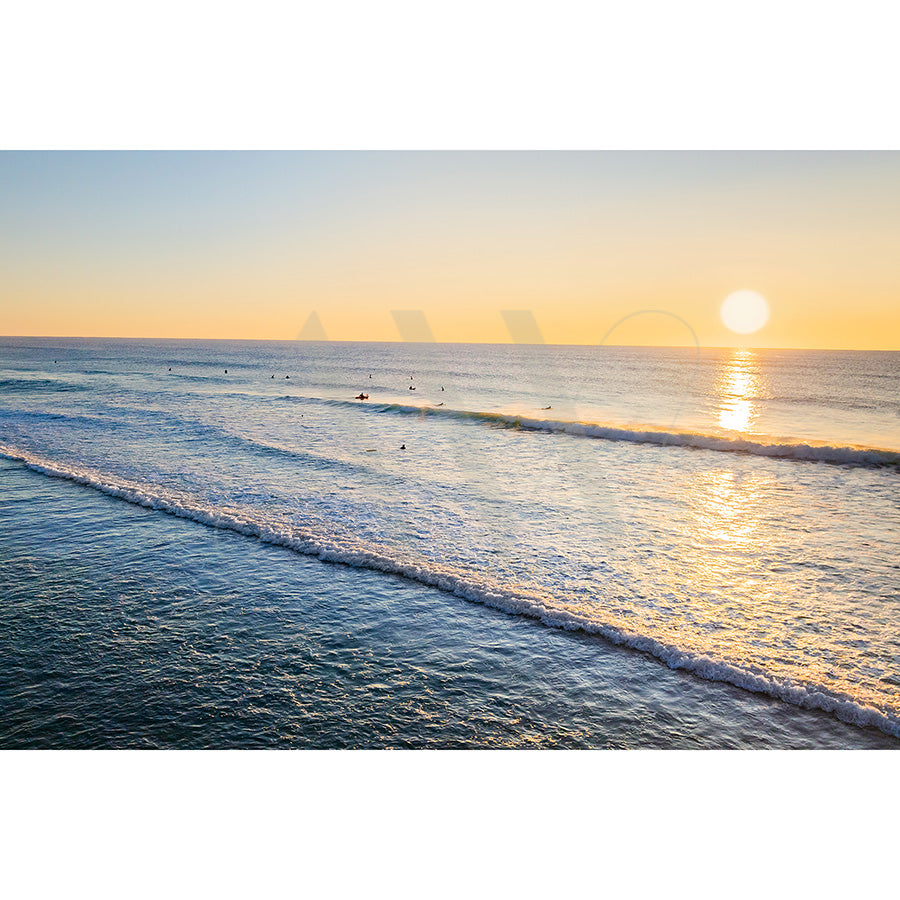 Peregian Beach Drone Image 0682 by AWP Image Library showcases a sunset over the ocean, with gentle waves and surfers; the clear sky allows the sun to cast a warm golden glow on the water, creating a tranquil and picturesque scene.