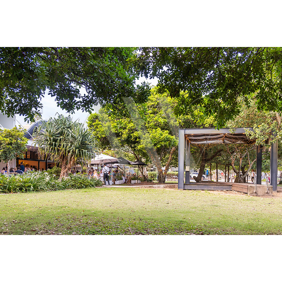 The Peregian Beach Village Image 2120 by AWP Image Library captures a peaceful, sunny park scene with lush green grass, trees, and a shelter structure on the right. People relax under umbrellas near a building on the left.
