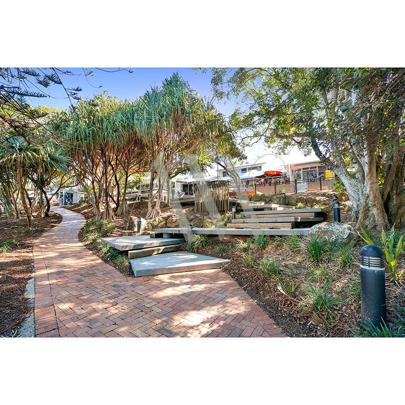 The Sunshine Beach Shops Image 2504 by AWP Image Library captures a scenic park with a brick-paved walkway surrounded by lush greenery and trees, featuring wooden benches alongside the path, with buildings visible in the backdrop beneath a clear blue sky.