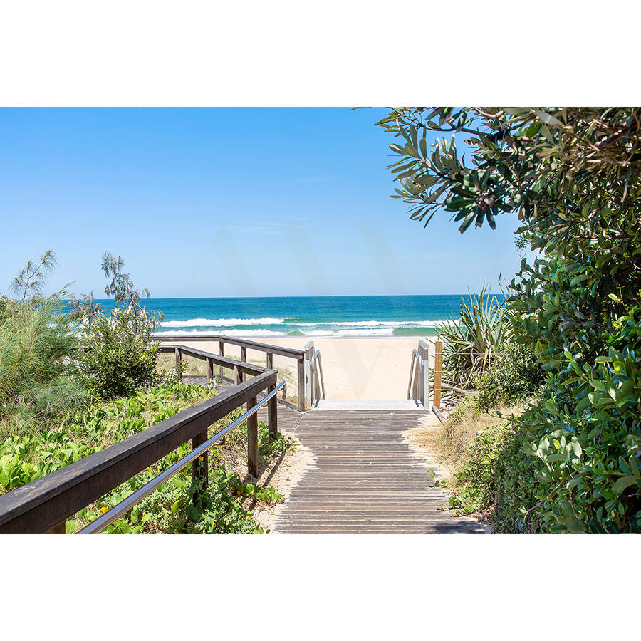 The Yaroomba Beach Image 3900 from AWP Image Library captures a wooden boardwalk winding through lush greenery to a sandy shore with gentle waves beneath a serene blue sky.