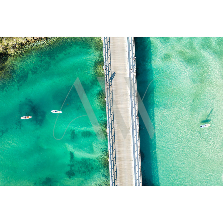 Brunswick Heads Drone Image 0197 from AWP Image Library captures an aerial view of a bridge over turquoise water. Three paddleboarders are on the left, while one is on the right, with the varying water shades creating a vibrant contrast around the bridge.