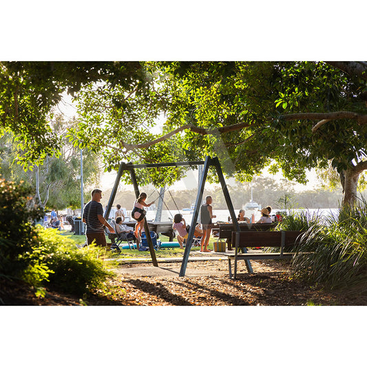 In Gympie Tce Image 3427 from AWP Image Library, children swing in a sunny park by the river, shaded by trees, while families relax on benches and grass, enjoying quality time as sunlight creates a lively atmosphere.