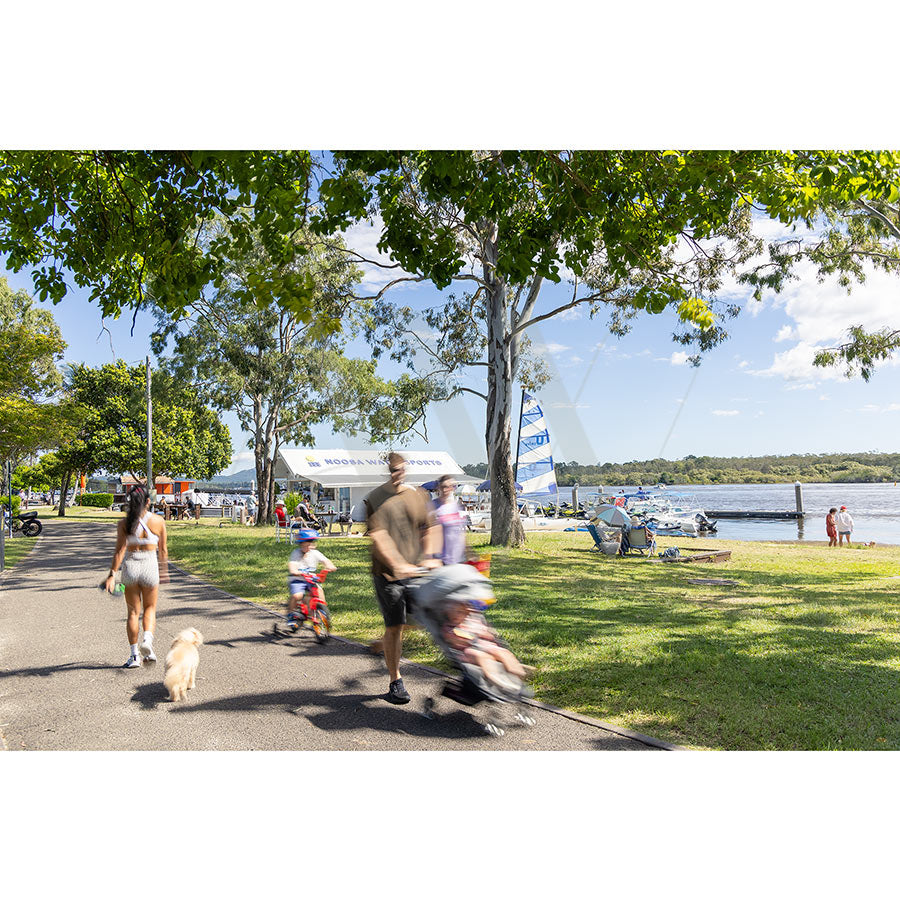 In Gympie Tce Image 4230 from AWP Image Library, people stroll and cycle on tree-lined paths while others unwind by the water, watching a Noosa Watersports sailboat glide beneath a bright, cloud-dotted sky.