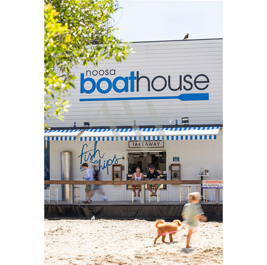 People relax outside Noosa Boathouse under a blue and white striped awning reading “fish & chips” in AWP Image Library’s Gympie Tce Image 4548, as a dog and child pass by on sandy ground near the Sunset Bar serving local fare.