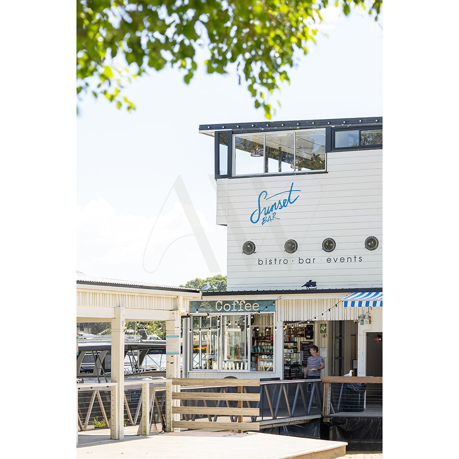 AWP Image Library’s “Gympie Tce Image 4596” shows Boathouse Noosa with Sunset Bar and Coffee signs, panoramic river views through large windows, porthole accents, a wooden deck, and local fare at the coffee counter beneath leafy trees.