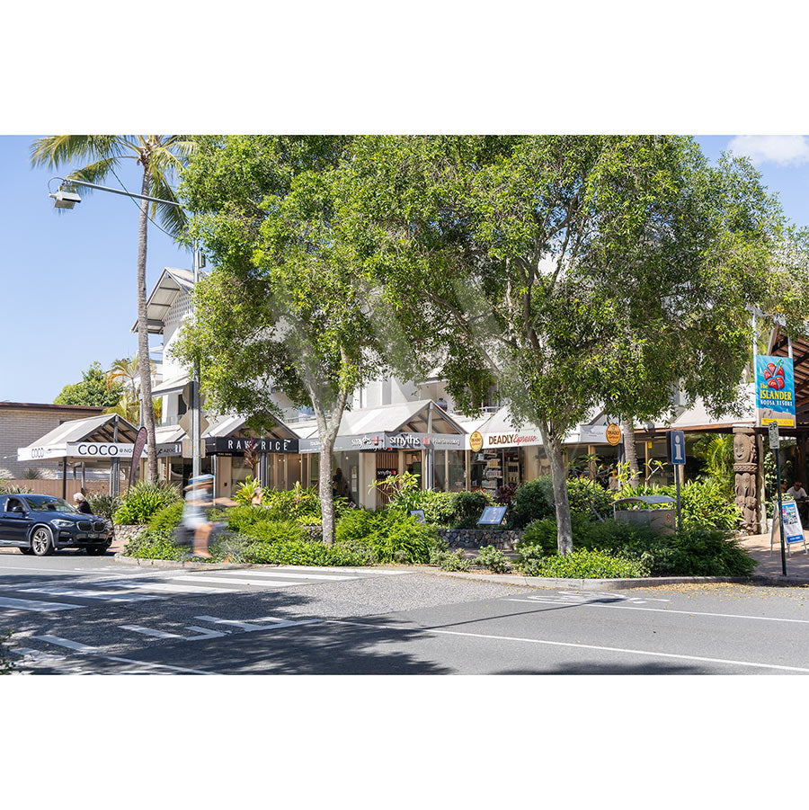 Gympie Tce Image 4745 by AWP Image Library shows shops with awnings along sunny Gympie Terrace, leafy trees, the Noosa River, business signs, a parked car, and a person walking under a clear blue sky.