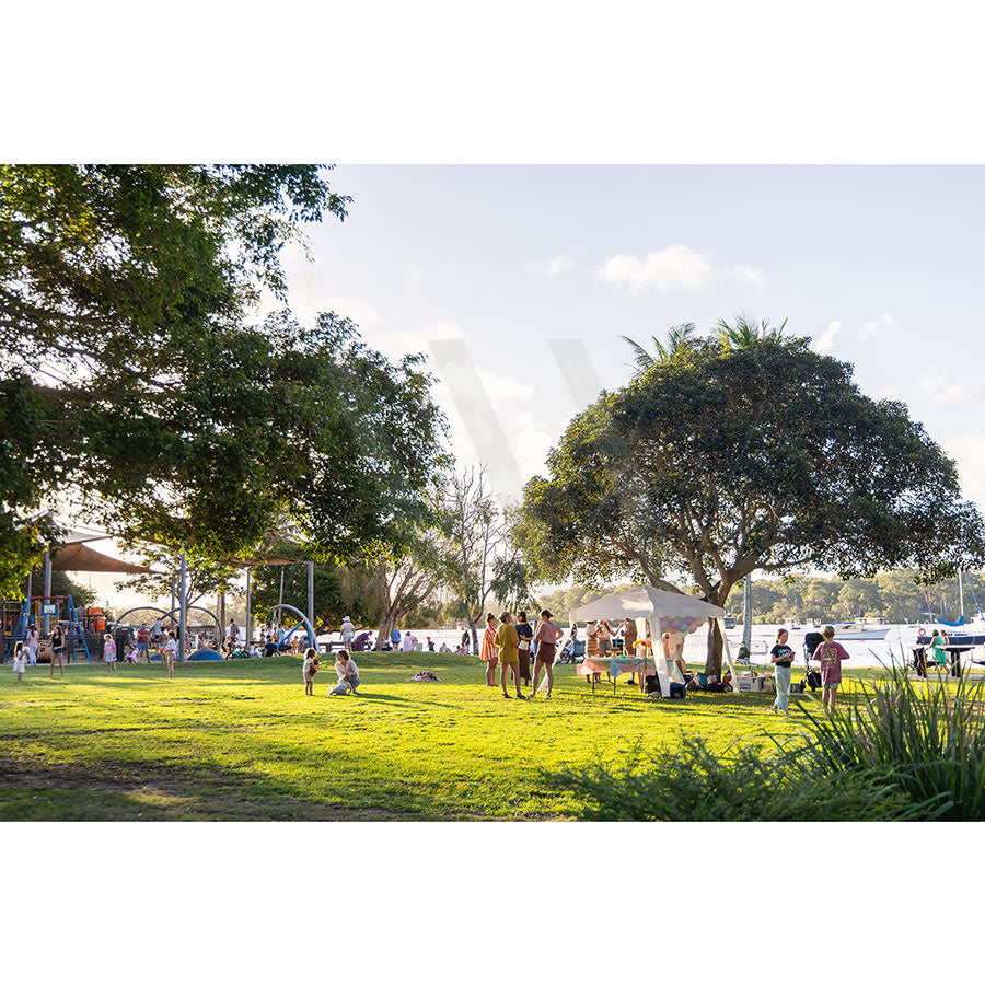 In Gympie Tce Image 3436 by AWP Image Library, people relax on lush grass under shady trees and a white tent in a sunny riverside park along the Noosa River, while others enjoy modern play equipment at the nearby Gympie Terrace playground.